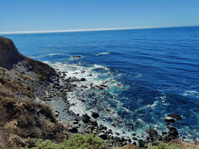 A rocky cove on the Big Sur coast with blue ocean and white surf below a scrubby bluff